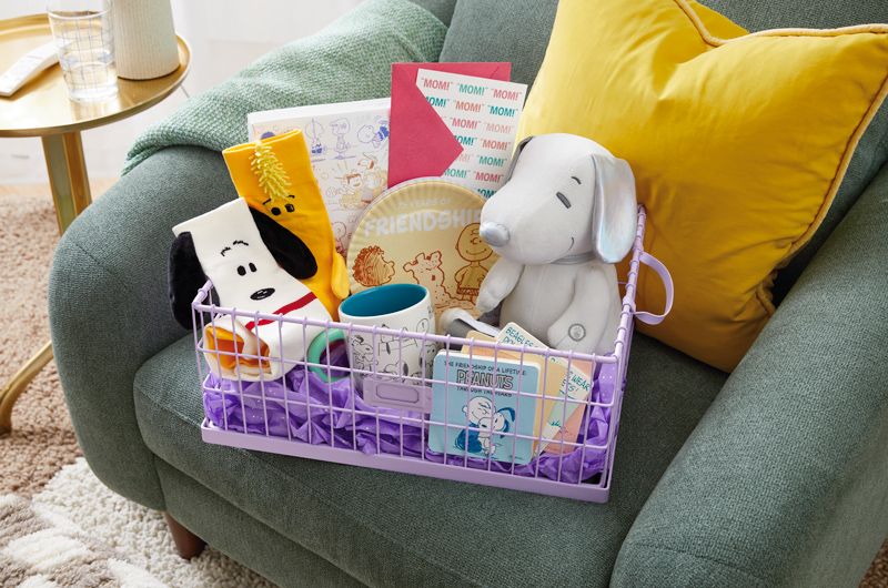 A Peanuts-themed Mother's Day gift basket that includes a pair of Snoopy and Woodstock socks, a mug with various illustrations of Snoopy on it, an interactive plush Snoopy sitting at a typewriter, a set of Peanuts snack plates, a set of coasters featuring scenes from the comics and a greeting card.